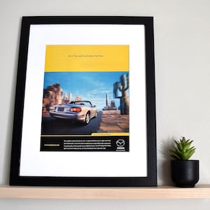 May include: Framed advertisement for the 1999 Mazda Miata. The image features a silver convertible car driving on a road with a desert landscape in the background. The top of the frame has yellow and blue sections with text. The frame is black.