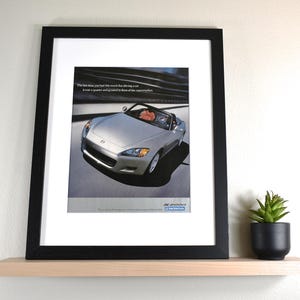 May include: Framed print of a silver Honda S2000 convertible. The image features the car in motion with a driver visible. Text at the top reads, "The last time you had this much fun driving a car..." The Honda logo is at the bottom.