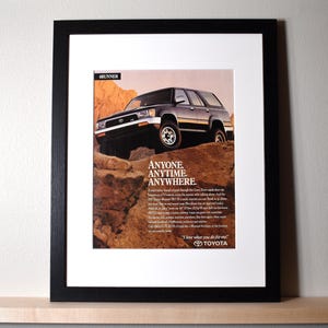 May include: Framed print featuring a black Toyota 4Runner SUV on a rocky terrain. The image includes the text "ANYONE. ANYTIME. ANYWHERE." and the Toyota logo. The print is in a black frame.
