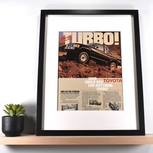 May include: Framed vintage advertisement for a black Toyota pickup truck, labeled "Turbo!" and "4x4." The ad features the truck driving on a dirt road. The text reads "Who could ask for anything more!" and includes specifications.