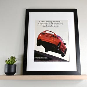 May include: Framed print featuring a red sports car with the text "It's not exactly a Ferrari. A Ferrari doesn't even have dual cup holders." The print is displayed on a light wood shelf with a small potted plant.