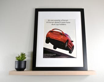 May include: Framed print featuring a red sports car with the text "It's not exactly a Ferrari. A Ferrari doesn't even have dual cup holders." The print is displayed on a light wood shelf with a small potted plant.