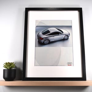May include: Framed print featuring a silver Audi TT sports car. The car is displayed on a white background with a subtle design element. The frame is black, and the print is mounted on a white mat. The Audi logo is visible.