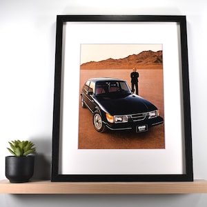 May include: Framed print featuring a black Saab 900 Turbo car on a desert landscape. The car is parked with a person standing nearby. The print is in a black frame and displayed on a wooden shelf with a small potted plant.