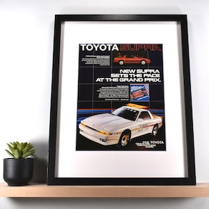 May include: Framed vintage Toyota Supra advertisement. The black framed print features a white car with the text "PACE CAR" and the Toyota logo. The ad includes the text "NEW SUPRA SETS THE PACE AT THE GRAND PRIX."