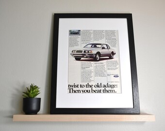May include: Framed vintage advertisement for a Ford Thunderbird. The ad features a silver car with text that reads "twist to the old adage: Then you beat them." The framed print is displayed on a light wood shelf with a small potted plant.