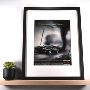 May include: Framed advertisement for the 2000 Chevy Monte Carlo. The image features a black car driving on a road with a tornado in the background. The frame is black, and the print is displayed on a white mat. A small potted succulent sits on a wooden shelf.