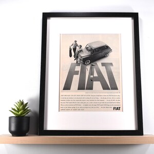 May include: Framed vintage Fiat advertisement. The black and white print features a classic Fiat car with two figures and the bold word "FIAT." The frame is black, and the print is displayed on a light-colored shelf with a small potted plant.