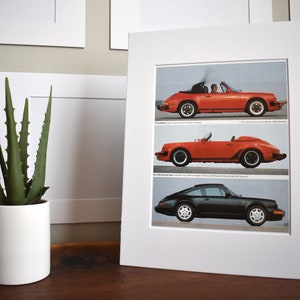 May include: A framed print of three different Porsche 911 models. The top image shows a red convertible with a driver in the car. The middle image shows a red convertible with the top down. The bottom image shows a black coupe.