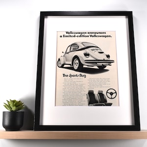 May include: Framed vintage advertisement for a limited-edition Volkswagen Beetle, the "Sport Bug." The black and white ad features the car's rear view, interior details, and text. The framed print sits on a wooden shelf with a small potted plant.