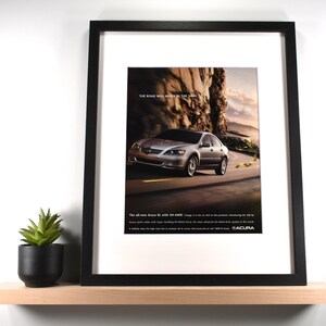 May include: Framed print featuring a silver Acura RL car driving on a winding road. The image has the text "THE ROAD WILL NEVER BE THE SAME." The print is in a black frame and is displayed on a wooden shelf with a small potted plant.