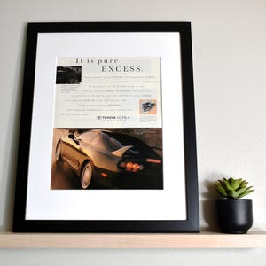 May include: Framed Toyota Supra advertisement. The black framed print features a car image and text that reads "It is pure EXCESS." The print is displayed on a light wood shelf with a small potted plant.