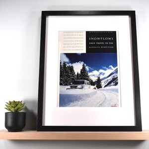 May include: Framed print featuring a snowplow on a snowy mountain road. The print has a black frame and white matting. The text on the print reads "SNOWPLOWS ONLY TRAVEL IN THE OPPOSITE DIRECTION."