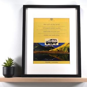 May include: Framed Toyota advertisement featuring a silver SUV against a backdrop of mountains and water. The ad includes the text "the call of the wild whispers your name, and asks if you can come out and play." The frame is black.
