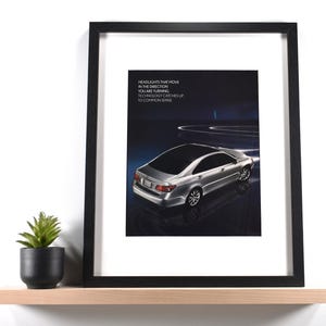 May include: Framed print featuring a silver car with the text "HEADLIGHTS THAT MOVE IN THE DIRECTION YOU ARE TURNING. TECHNOLOGY CATCHES UP TO COMMON SENSE." The print is in a black frame, displayed on a wooden shelf with a small potted plant.