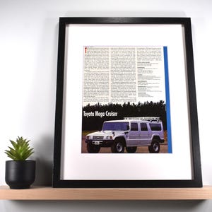 May include: Framed print featuring a vintage advertisement for the Toyota Mega Cruiser. The ad showcases a silver SUV with a roof rack against a backdrop of trees. The text "Toyota Mega Cruiser" is prominently displayed.