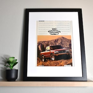 May include: Framed print of a red Mazda pickup truck driving on a dirt road in a mountainous landscape. The print includes the text "100% Mountain Grown" and the Mazda logo. The frame is black, and the print is displayed on a wooden shelf.