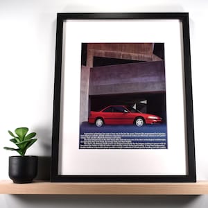 May include: Framed print of a red Acura Integra car ad. The car is parked under a concrete structure. The print is in a black frame and sits on a wooden shelf with a small potted plant.