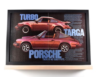 May include: Framed vintage advertisement featuring two red Porsche cars, a Turbo and a Targa model. The advertisement has the words "TURBO", "TARGA", and "PORSCHE" in blue text. The frame is black and sits on a light-colored wooden shelf.