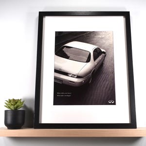 May include: Framed black and white print of a white car, likely an Infiniti, on a road. The print is in a black frame with a white mat. The Infiniti logo is visible. Text at the bottom reads, "What inspires your heart? What makes you happy?"
