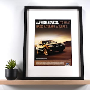 May include: Framed Subaru advertisement featuring a black SUV driving on a road. The ad includes the text "ALL-WHEEL REFLEXES. IT'S WHAT MAKES A SUBARU, A SUBARU." The frame is black, and the print is displayed on a wooden shelf with a small potted plant.
