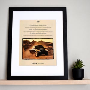 May include: Framed print featuring a Toyota truck ad. The ad shows a black truck driving through a dusty landscape. Text at the top reads "Goats understand your need to climb mountains." The Toyota logo is at the bottom.