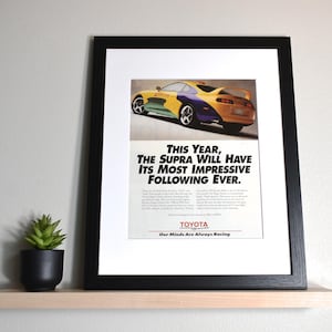 May include: Framed Toyota Supra advertisement. The image features a colorful sports car with text that reads "THIS YEAR, THE SUPRA WILL HAVE ITS MOST IMPRESSIVE FOLLOWING EVER." The frame is black and sits on a wooden shelf.