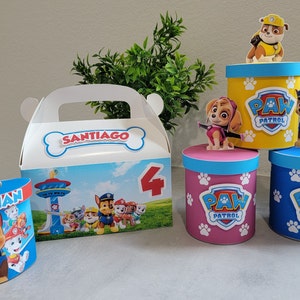 May include: Four Paw Patrol themed party favor boxes. Two boxes are round with blue and white paw prints and a Paw Patrol character on top. One box is round with pink and white paw prints and a Paw Patrol character on top. One box is a rectangular take-out style box with a Paw Patrol character on the front and the name Santiago and the number 4.