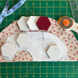 May include: A quilting project in progress, featuring a floral fabric, white and burgundy hexagon fabric pieces, a rotary cutter, and a measuring tape. The project is on a green cutting mat.