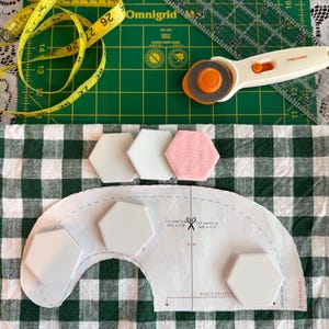 May include: A sewing project in progress, featuring a green cutting mat, rotary cutter, and measuring tape. White and pink hexagon-shaped fabric pieces are arranged on a green and white checkered fabric, along with a pattern piece.