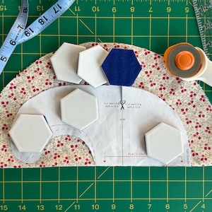 May include: A quilting project in progress, featuring a floral fabric, white and blue hexagonal tiles, and a paper pattern. A rotary cutter and measuring tape are also present on the green cutting mat.