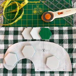 May include: A sewing project in progress, featuring a green cutting mat, a yellow measuring tape, and a rotary cutter. White and green hexagon-shaped pieces are arranged on a green and white checkered fabric, along with a pattern.