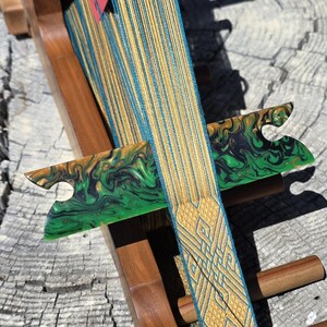 May include: A wooden inkle loom holding a warp of multicolored threads in shades of blue, orange, and yellow. The loom features two green and black marbled resin heddle sticks and a woven band with a geometric pattern extending from the front.