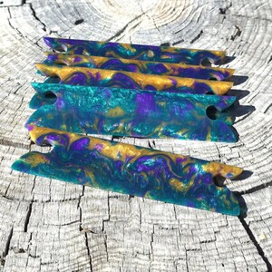 May include: A set of four rectangular resin yarn bobbins featuring a marbled swirl pattern in shades of metallic gold, deep purple, and teal green. Each bobbin has a notched cutout on both ends to secure yarn, with a smooth, glossy finish.