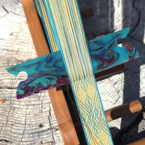 May include: A wooden inkle loom holding a warp of teal and yellow threads. A flat, marbled teal and purple shuttle rests horizontally across the tensioned threads, which feature a complex woven geometric pattern in yellow and teal.