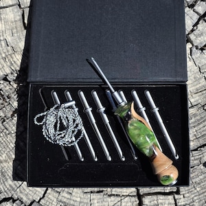 May include: A set of interchangeable crochet hooks with a decorative handle made of wood and green resin, housed in a black presentation box. The set includes multiple metal hook tips, a silver cord, and a single handle with a marbled green finish.