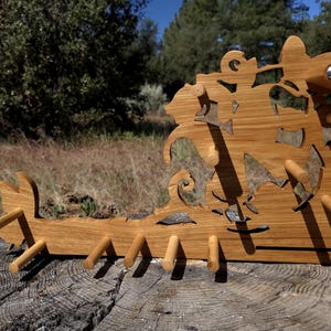 Puede incluir: Un perchero de madera con un diseño artístico único. El perchero está hecho de madera de color claro y presenta formas recortadas y varias clavijas para colgar objetos. Se coloca al aire libre sobre un tocón de árbol, con un fondo natural de árboles y follaje.