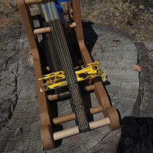 Puede incluir: Un telar de madera con hilos negros, amarillos y azules. El telar tiene una pieza decorativa con estampado amarillo y negro y una banda tejida con un diseño geométrico. El telar está sobre un tocón de árbol.