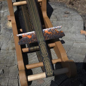 Puede incluir: Un telar de madera con hilos negros, verdes y dorados. La lanzadera tiene un diseño veteado en naranja, blanco y negro. La banda tejida presenta un patrón geométrico en oro y verde oscuro.