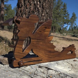 Puede incluir: Un soporte de madera marrón oscuro con forma de hada reclinada con alas. El objeto presenta una base horizontal, una estructura de soporte vertical y dos pequeños pomos de madera con tornillos visibles fijados en la parte frontal.