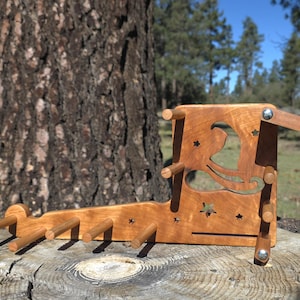 May include: A wooden yarn swift and ball winder, crafted from light-colored wood. The swift features a decorative cutout design of a witch and stars. The ball winder has several pegs for holding yarn. The item is displayed outdoors on a tree stump.