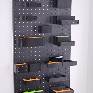 Può includere: Un organizzatore da parete in bacheca nera con vari contenitori di stoccaggio in plastica nera. Alcuni contenitori hanno accenti arancioni e verdi. I contenitori sono di diverse dimensioni e forme, tra cui rettangolari, quadrati e triangolari.