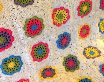 May include: A white crocheted blanket with colorful flower motifs. The flowers are made with different shades of pink, yellow, green, and blue yarn.