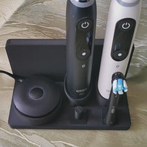 Könnte beinhalten: Eine schwarze Ladestation mit zwei elektrischen Zahnbürsten und einer Ladebasis. Eine Zahnbürste ist schwarz, die andere weiß. Die Ladebasis ist rund und hat einen kleinen, zylindrischen Pfosten.