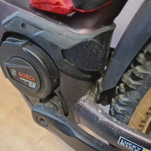 Pode incluir: Close-up de um motor de bicicleta elétrica preto e cinza com o texto "BOSCH Performance CX". A imagem também mostra uma parte do pneu e da estrutura. A bicicleta tem um guarda-lamas preto e uma bolsa vermelha.