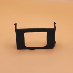 Może przedstawiać: Czarny plastikowy element z prostokątnym otworem. Element ma górną część w kształcie litery U i małe wypukłości po prawej stronie. Tło jest w jednolitym kolorze beżowym.