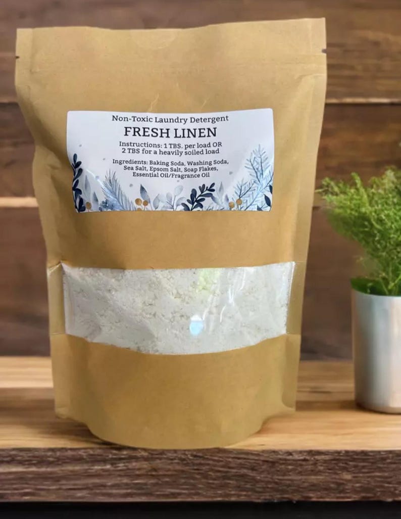 May include: A kraft paper bag of "Fresh Linen" non-toxic laundry detergent. The bag has a clear window showing the white powder inside. The label includes instructions and ingredients: Baking Soda, Washing Soda, Sea Salt, Epsom Salt, Soap Flakes, and Essential Oil/Fragrance Oil.