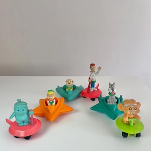 May include: A collection of colorful, vintage toy figures in miniature flying saucers. The figures include cartoon characters in various colors, such as blue, orange, and green. The toys are on a white surface.