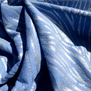 Op de afbeelding: Blauwe stof met een zilveren metallic patroon. De stof heeft een textuur en is gevouwen om het patroon te laten zien.