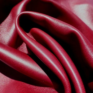 Puede incluir: Primer plano de tela de cuero rojo oscuro con una textura sutil. El material está doblado y drapeado, creando curvas suaves y reflejos. El color es un tono rico y saturado, con un ligero brillo.